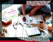 A person sits cross-legged on a wooden floor assembling a vision board, surrounded by scattered photos, paper, markers, a notebook, glasses, a mug, and a lit candle, with hands arranging images and writing on a sheet.